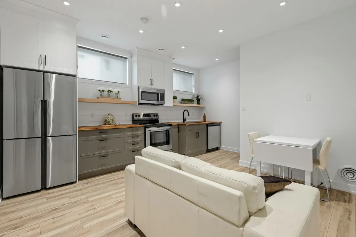 Phòng khách liền bếp chữ L, sofa xám và cửa kính mở ra ban công có chậu cây